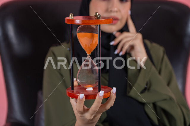 The importance of punctuality and commitment to appointments, close-up of an hourglass, business management and schedule organization, monitoring sand timing, realizing the value of precious moments, portrait of a veiled Emirati Gulf Arab woman wearing a green abaya sitting on a comfortable chair and holding a bank card, pink background