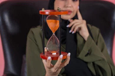 The importance of punctuality and commitment to appointments, close-up of an hourglass, business management and schedule organization, monitoring sand timing, realizing the value of precious moments, portrait of a veiled Emirati Gulf Arab woman wearing a green abaya sitting on a comfortable chair and holding a bank card, pink background