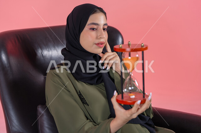 القيام بمراقبة توقيت الرمل, مفهوم إدارة الأعمال وتنظيم الجدول الزمني, إدراك قيمة اللحظات الثمينة,  بورتريه لامرأة اماراتية خليجية عربية محجبة ترتدي عباءة خضراء تجلس على كرسي مريح و تمسك ساعة رملية وتضع يدها على خدها, التحليل والتدقيق واتخاذ القرارات, مفهوم التمعن والتركيز بالوقت, خلفية وردية