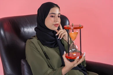 القيام بمراقبة توقيت الرمل, مفهوم إدارة الأعمال وتنظيم الجدول الزمني, إدراك قيمة اللحظات الثمينة,  بورتريه لامرأة اماراتية خليجية عربية محجبة ترتدي عباءة خضراء تجلس على كرسي مريح و تمسك ساعة رملية وتضع يدها على خدها, التحليل والتدقيق واتخاذ القرارات, مفهوم التمعن والتركيز بالوقت, خلفية وردية