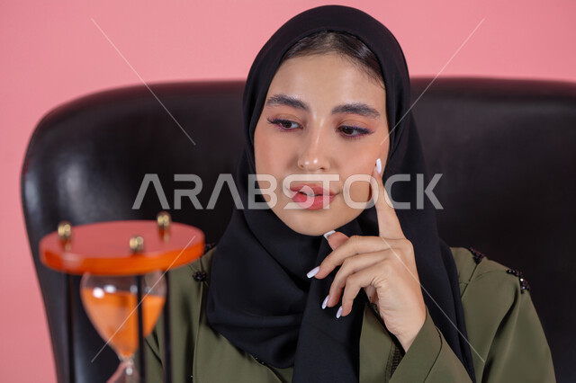 Analysis and decision making, thinking about the past and the future, the concept of contemplation and focusing on time, a portrait of a veiled Arab Emirati Gulf woman wearing a green abaya sitting on a comfortable chair holding an hourglass and placing her hand on her cheek, remembering memories and beautiful situations, pink background