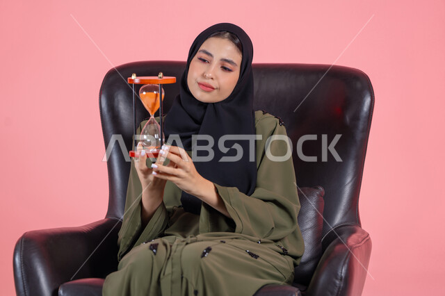 Deep mental thinking, thinking about the past and the future, the concept of contemplating and focusing on time, realizing the value of precious moments, portrait of a veiled Arab Emirati Gulf woman wearing a green abaya sitting on a comfortable chair and holding an hourglass, pink background