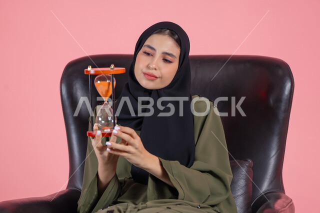 الجلوس بتعابير الملل والانتظار و السؤال, إدارة الأعمال و تنظيم الجدول الزمني, القيام بمراقبة توقيت الرمل, إدراك قيمة اللحظات الثمينة,  بورتريه لامرأة اماراتية خليجية عربية محجبة ترتدي عباءة خضراء تجلس على كرسي مريح و تمسك ساعة رملية, خلفية وردية
