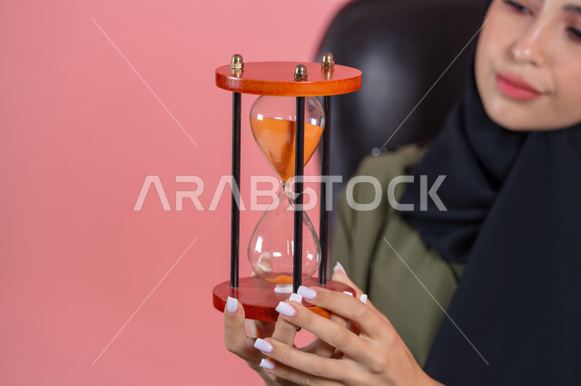 صوره مقربه لساعه رمليه,  إدارة الأعمال و تنظيم الجدول الزمني, القيام بمراقبة توقيت الرمل, إدراك قيمة اللحظات الثمينة, بورتريه لامرأة اماراتية خليجية عربية محجبة ترتدي عباءة خضراء تجلس على كرسي مريح و تمسك البنكام , خلفية وردية