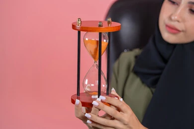 صوره مقربه لساعه رمليه,  إدارة الأعمال و تنظيم الجدول الزمني, القيام بمراقبة توقيت الرمل, إدراك قيمة اللحظات الثمينة, بورتريه لامرأة اماراتية خليجية عربية محجبة ترتدي عباءة خضراء تجلس على كرسي مريح و تمسك البنكام , خلفية وردية