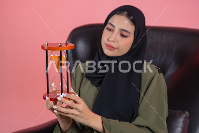 أدوات قديمة لتحديد الوقت، الجلوس بتعابير الملل والانتظار و السؤال, إدارة الأعمال و تنظيم الجدول الزمني, القيام بمراقبة توقيت الرمل, إدراك قيمة اللحظات الثمينة, بورتريه لامرأة اماراتية خليجية عربية محجبة ترتدي عباءة خضراء تجلس على كرسي مريح و تمسك ساعة رملية, خلفية وردية