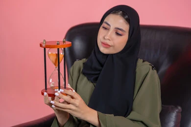 Old tools for telling time, sitting with bored expressions, waiting and asking, business management and organizing schedule, monitoring sand timing, realizing the value of precious moments, portrait of a veiled Arab Gulf Emirati woman wearing a green abaya sitting on a comfortable chair and holding an hourglass, pink background