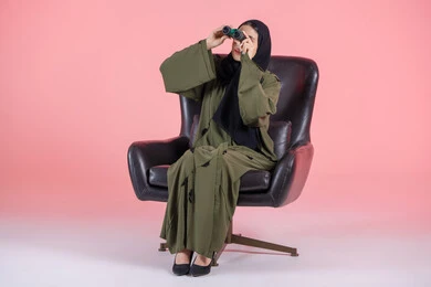 رسم خطة مستقبلية، رؤية الاشياء البعيدة و تقريبها عبر التليكسوب، تعابير الاندماج والجدية, بورتريه  لامرأة اماراتية عربية خليجية محجبة ترتدي العباءة تجلس على كرسي تحمل بيديها منظار ثنائي العينين و تنظر الى شئ ما, خلفية وردية