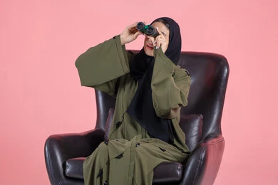 Setting future goals, seeing distant things and bringing them closer through a telescope, expressions of integration and seriousness, a portrait of an Emirati Arab Gulf woman wearing a hijab and an abaya sitting on a chair holding binoculars in her hands and looking at something, pink background.