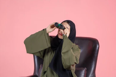 التخطيط بعناية للمستقبل، رؤية الاشياء البعيدة و تقريبها عبر التليكسوب، تعابير الاندماج والجدية, بورتريه  لامرأة اماراتية عربية خليجية محجبة ترتدي العباءة تجلس على كرسي تحمل بيديها منظار ثنائي العينين و تنظر الى شئ ما, خلفية وردية