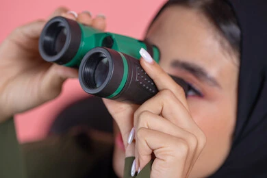 تعابير الاندماج والجدية، التخطيط المستقبلي للأهداف والطموحات، رؤية الاشياء البعيدة عبر التلسكوب, بورتريه  مقرب لامرأة اماراتية عربية خليجية محجبة ترتدي العباءة تحمل بيديها منظار ثنائي العينين و تنظر الى شئ ما, خلفية وردية