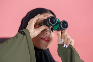 التخطيط المستقبلي للأهداف والطموحات، تعابير الاندماج والجدية، رؤية الاشياء البعيدة عبر التلسكوب, بورتريه  مقرب لامرأة اماراتية عربية خليجية محجبة ترتدي العباءة تحمل بيديها منظار ثنائي العينين و تنظر الى شئ ما, خلفية وردية