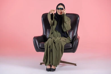 The careful observation of distant things, a love for exploration and adventure, a portrait of an Emirati Arab Gulf woman wearing a hijab and an abaya sitting on a chair holding binoculars, pointing with her index finger at something, future planning for goals and ambitions, pink background.