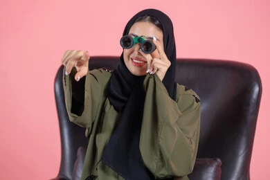 Looking at distant horizons, a love for exploration and adventure, a close-up portrait of an Emirati Arab Gulf woman wearing a hijab and an abaya sitting on a chair holding binoculars, pointing with her index finger at something, future planning for goals and ambitions, pink background.