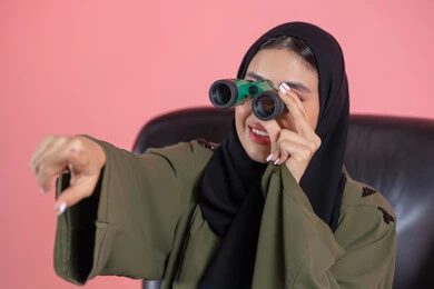 Observing distant things, a love for exploration and adventure, a close-up portrait of an Emirati Arab Gulf woman wearing a hijab and an abaya sitting on a chair holding binoculars, pointing with her index finger at something, future planning for goals and ambitions, pink background.