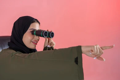 Seeing distant things, a love for exploration and adventure, a close-up portrait of an Emirati Arab Gulf woman wearing a hijab and an abaya sitting on a chair holding binoculars, pointing with her index finger at something, future planning for goals and ambitions, pink background.