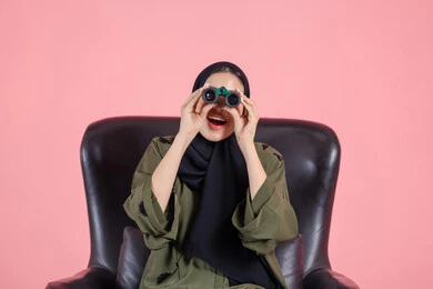 شغف الاكتشاف وخوض المغامرات، ايماءات الاستمتاع و السرور، رؤية الاشياء البعيدة عبر التلسكوب, بورتريه مقرب لامرأة اماراتية عربية خليجية محجبة ترتدي العباءة تجلس على كرسي تحمل بيديها منظار ثنائي العينين و تنظر الى شئ ما, خلفية وردية