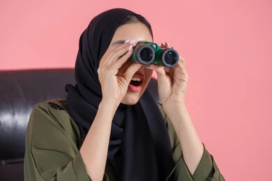 ايماءات الاستمتاع و السرور، حب الاستكشاف و المغامرة، رؤية الاشياء البعيدة عبر التلسكوب, بورتريه مقرب لامرأة اماراتية عربية خليجية محجبة ترتدي العباءة تحمل بيديها منظار ثنائي العينين و تنظر الى شئ ما, خلفية وردية