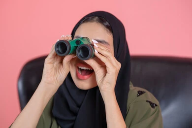 حب الاستكشاف و المغامرة، رؤية الاشياء البعيدة عبر التلسكوب, بورتريه مقرب لامرأة اماراتية عربية خليجية محجبة ترتدي العباءة تحمل بيديها منظار ثنائي العينين و تنظر الى شئ ما, ايماءات الاستمتاع و السرور, خلفية وردية