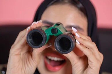 Seeing distant things through a telescope, a love for exploration and adventure, a close-up portrait of an Emirati Arab Gulf woman wearing a hijab and an abaya, holding binoculars in her hands and looking at something, gestures of enjoyment and pleasure, pink background.