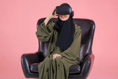 Spending enjoyable time playing and having fun, enjoying watching virtual reality movies, interacting and immersing oneself in a simulated fantasy world, a portrait of an Emirati Arab Gulf woman wearing a hijab and an abaya, with 3D VR glasses, sitting on a chair looking intently at something, with a pink background.