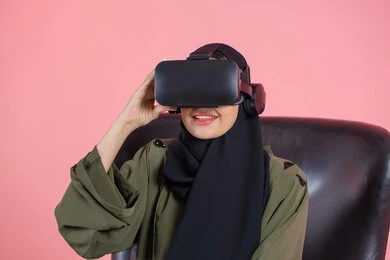 الترفيه والتسلية في وقت الفراغ، الاستمتاع بمشاهدة أفلام الواقع الافتراضي، التفاعل والاندماج ومحاكاة العالم الخيالي, بورتريه مقرب لامرأة اماراتية عربية خليجية محجبة ترتدي العباءة ونظارة في آر ثلاثية الأبعاد تجلس على كرسي تنظر إلى شيء ما بتركيز, خلفية وردية
