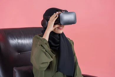 التفاعل والاندماج ومحاكاة العالم الخيالي, الاستمتاع بمشاهدة أفلام الواقع الافتراضي, بورتريه لامرأة اماراتية عربية خليجية محجبة ترتدي العباءة ونظارة في آر ثلاثية الأبعاد تجلس على الكرسي وتنظر إلى شيء ما بتركيز, الترفيه والتسلية في وقت الفراغ, خلفية وردية