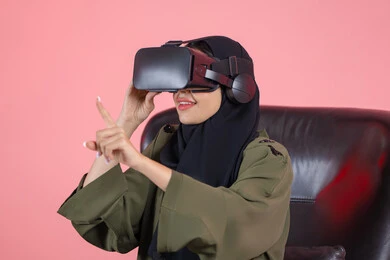 إيماءات التفاعل والاندماج، مفهوم التقنيات الإلكترونية الحديثة، بورتريه مقرب لامرأة اماراتية عربية خليجية محجبة ترتدي العباءة و نظارات الواقع الإفتراضي (في آر) ثلاثية الأبعاد تجلس على كرسي تقوم بالنقرعلى شيء ما، خلفية ملونة