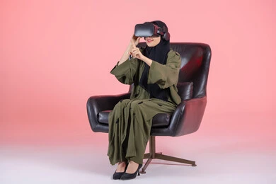 The concept of modern electronic technologies, interaction and integration gestures, a portrait of an Emirati Arab Gulf woman wearing a hijab and an abaya, wearing 3D virtual reality (VR) glasses, sitting on a chair and clicking on something, colorful background.
