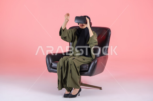 المتعة بالمشاهدة عبر نظارة الواقع الافتراضي، دمج التسلية مع العالم الخيالي، رفع كف اليد للأعلى بإيماءات الاندماج، التطور التقني والتكنولوجي, بورتريه لامرأة اماراتية عربية خليجية محجبة ترتدي عباءة خضراء تجلس على كرسي مريح و تضع نظارة الفي ار, خلفية وردية