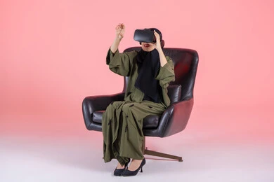 المتعة بالمشاهدة عبر نظارة الواقع الافتراضي، دمج التسلية مع العالم الخيالي، رفع كف اليد للأعلى بإيماءات الاندماج، التطور التقني والتكنولوجي, بورتريه لامرأة اماراتية عربية خليجية محجبة ترتدي عباءة خضراء تجلس على كرسي مريح و تضع نظارة الفي ار, خلفية وردية