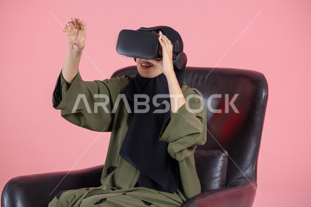 Merging entertainment with the imaginary world, raising the palm of the hand upwards with gestures of integration and simulation of the imaginary world, enjoyment of watching through virtual reality glasses, technical and technological development, close-up portrait of a veiled Emirati Arab Gulf woman wearing a green abaya and VR glasses, pink background