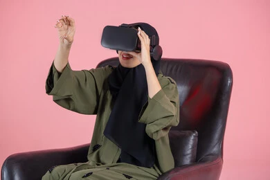 دمج التسلية مع العالم الخيالي، رفع كف اليد للأعلى بإيماءات الاندماج ومحاكاة العالم الخيالي، المتعة بالمشاهدة عبر نظارة الواقع الافتراضي، التطور التقني والتكنولوجي, بورتريه مقرب لامرأة اماراتية عربية خليجية محجبة ترتدي عباءة خضراء و تضع نظارة الفي ار, خلفية وردية