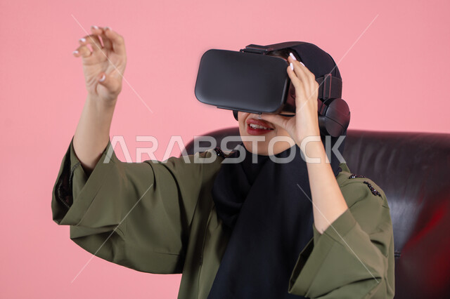 Raising the palm of the hand up with gestures of integration and simulating the imaginary world, enjoying watching through virtual reality glasses, technical and technological development, close-up portrait of a veiled Emirati Arab Gulf woman wearing a green abaya and VR glasses, pink background