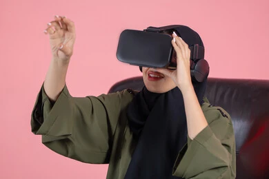 Raising the palm of the hand up with gestures of integration and simulating the imaginary world, enjoying watching through virtual reality glasses, technical and technological development, close-up portrait of a veiled Emirati Arab Gulf woman wearing a green abaya and VR glasses, pink background