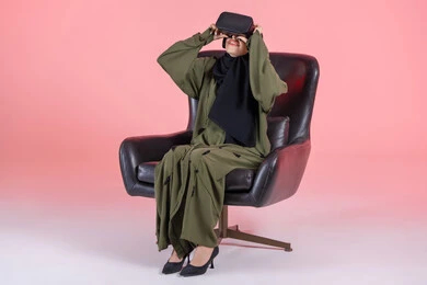 Integration and interaction with virtual reality, spending leisure time on recreational activities, technological and technical development, a portrait of a Gulf Arab Emirati woman wearing a hijab sitting on a chair wearing an abaya and 3D glasses with expressions of joy, pink background.