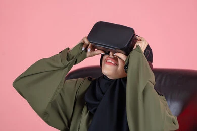 الاستمتاع بمشاهدة أفلام الواقع الافتراضي ومحاكاة العالم الخيالي، التطور التقني والتكنولوجي, قضاء وقت الفراغ بالنشاط الترفيهي, بورتريه مقرب لامرأة عربية خليجية اماراتية محجبة تجلس على كرسي ترتدي العباءة و نظارة في آر ثلاثية الأبعاد بتعابير السرور, خلفية وردية