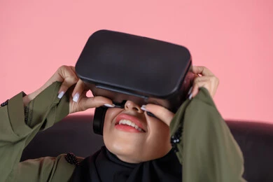 التطور التقني والتكنولوجي, الاستمتاع بمشاهدة أفلام الواقع الافتراضي ومحاكاة العالم الخيالي, قضاء وقت الفراغ بالنشاط الترفيهي, بورتريه مقرب لامرأة عربية خليجية اماراتية محجبة تجلس على كرسي ترتدي العباءة و نظارة في آر ثلاثية الأبعاد بتعابير السرور, خلفية وردية