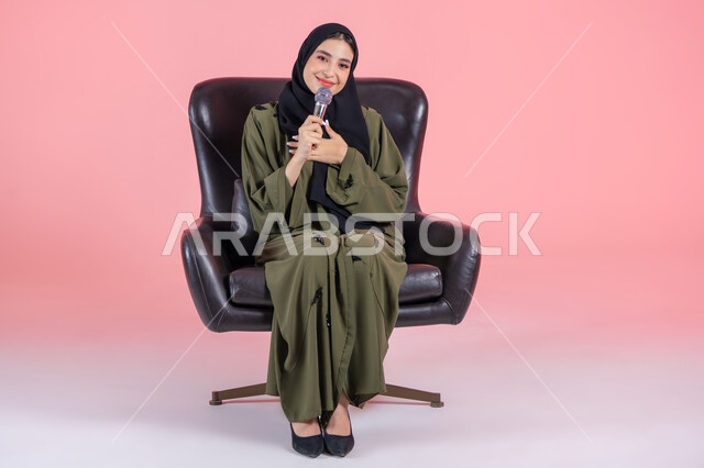 إجراء المقابلات وطرح الأسئلة، ايماءات الشكر و السعادة, لقاء الخطابات والندوات, تقديم العروض الفنيه و الغنائيه والفقرات الترفيهية، بدء التدريب الصوتي، مفهوم الفن وتأثيره الايجابي, بورتريه لامرأة اماراتية عربية خليجية محجبة مبتسمة ترتدي عباءة خضراء تجلس على كرسي مريح و تمسك بيدها المايكروفون, خلفية وردية