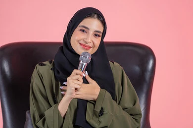 لقاء الخطابات والندوات, تقديم العروض الفنيه و الغنائيه والفقرات الترفيهية، بدء التدريب الصوتي، مفهوم الفن وتأثيره الايجابي, بورتريه لامرأة اماراتية عربية خليجية محجبة مبتسمة            ترتدي عباءة خضراء تجلس على كرسي مريح و تمسك بيدها المايكروفون, ايماءات الشكر والسعادة, خلفية وردية