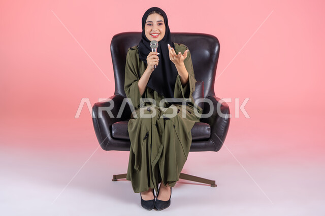 تقديم العروض الفنيه و الغنائيه والفقرات الترفيهية، بدء التدريب الصوتي، مفهوم الفن وتأثيره الايجابي, بورتريه لامرأة اماراتية عربية خليجية محجبة ترتدي عباءة خضراء تجلس على كرسي مريح و تمسك بيدها المايكروفون بإيماءات الاندماج بالغناء، مهارة إلقاء الخطابات والندوات، خلفية وردية
