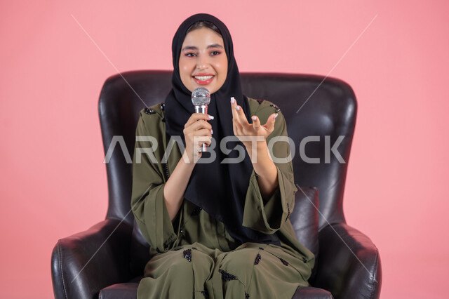 بدء التدريب الصوتي، تقديم العروض الفنيه و الغنائيه والفقرات الترفيهية، مفهوم الفن وتأثيره الايجابي, بورتريه لامرأة اماراتية عربية خليجية محجبة ترتدي عباءة خضراء تجلس على كرسي مريح و تمسك بيدها المايكروفون بإيماءات الاندماج بالغناء، مهارة إلقاء الخطابات والندوات، خلفية وردية