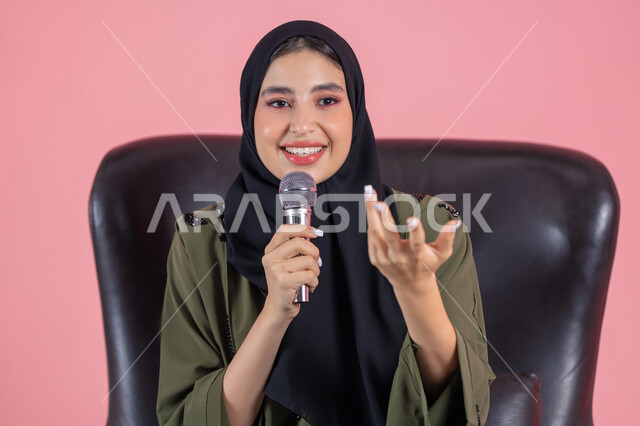 Discussion and effective communication, the art of public speaking and oratory, presenting artistic and singing performances and entertainment segments, the concept of art and its positive impact, a portrait of a veiled Emirati Arab Gulf woman wearing a green abaya sitting on a comfortable chair and holding a microphone in her hand with gestures of integration and happiness, pink background