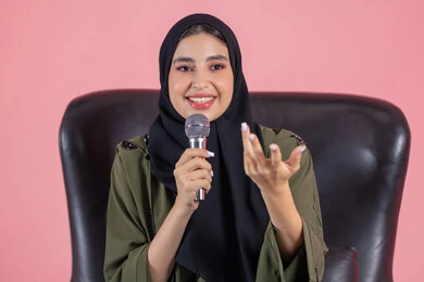 النقاش والتواصل الفعال، فن الالقاء والخطابة،  تقديم العروض الفنيه و الغنائيه والفقرات الترفيهية، مفهوم الفن وتأثيره الايجابي, بورتريه لامرأة اماراتية عربية خليجية محجبة ترتدي عباءة خضراء تجلس على كرسي مريح و تمسك بيدها المايكروفون بإيماءات الاندماج و السعادة, خلفية وردية