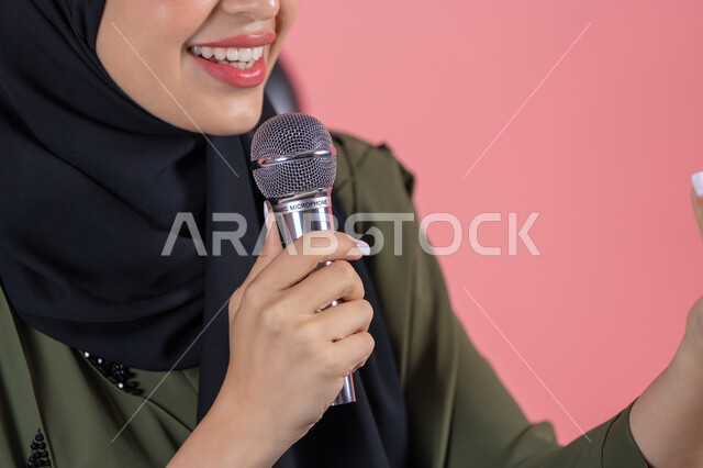 Vocal training and performance concept, recording singing clips, close-up portrait of a smiling Emirati Gulf Arab woman wearing a green abaya speaking into a microphone, presenting artistic and singing performances and entertainment segments, the concept of art and its positive impact, pink background