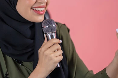 مفهوم التدريب والآداء الصوتي، تسجيل مقاطع غنائية، بورتريه مقرب لامرأة عربية خليجية اماراتية مبتسمة ترتدي عباءة خضراء  تتحدث عبر المايكروفون,  تقديم العروض الفنيه و الغنائيه والفقرات الترفيهية، مفهوم الفن وتأثيره الايجابي, خلفية وردية