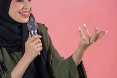 The art of giving speeches and seminars, a close-up portrait of a young Arab Gulf Emirati woman wearing a hijab and an abaya, holding a microphone in her hand, voice training and program presentation, pink background.