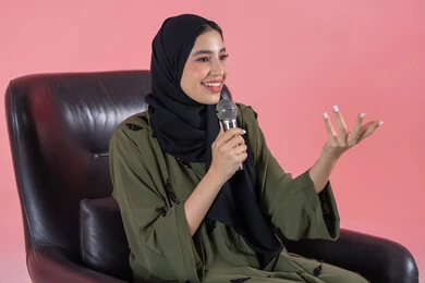 Speaking and dialogue skills, the art of elocution and rhetoric, starting vocal training, presenting artistic and singing performances and entertainment segments, the concept of art and its positive impact, a portrait of a veiled Emirati Arab Gulf woman wearing a green abaya sitting on a comfortable chair, holding a microphone and raising her hand with gestures of integration and focus, pink background