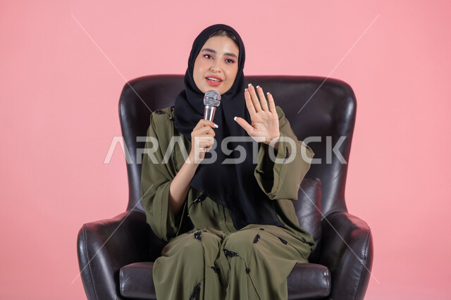 Voice training and radio program presentation, discussion and effective communication, the art of public speaking and oratory, presenting artistic and singing performances and entertainment segments, the concept of art and its positive impact, a portrait of a veiled Emirati Arab Gulf woman wearing a green abaya sitting on a comfortable chair and holding a microphone in her hand with gestures of integration and happiness, pink background