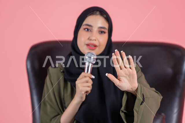 تقديم العروضات المسرحية، الاستمتاع بأداء المواهب الصوتية، مفهوم الفن وتأثيره الايجابي, بورتريه لامرأة اماراتية عربية خليجية محجبة ترتدي عباءة خضراء تجلس على كرسي مريح و تمسك المايكروفون و ترفع يدها بإيماءات الاندماج والتركيز، خلفية وردية